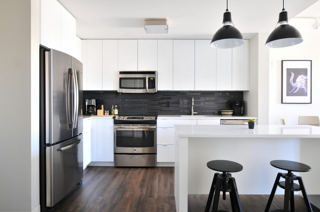 Modern white kitchen with stainless steel appliances, black tile backsplash, pendant lighting, and minimalist island seating.