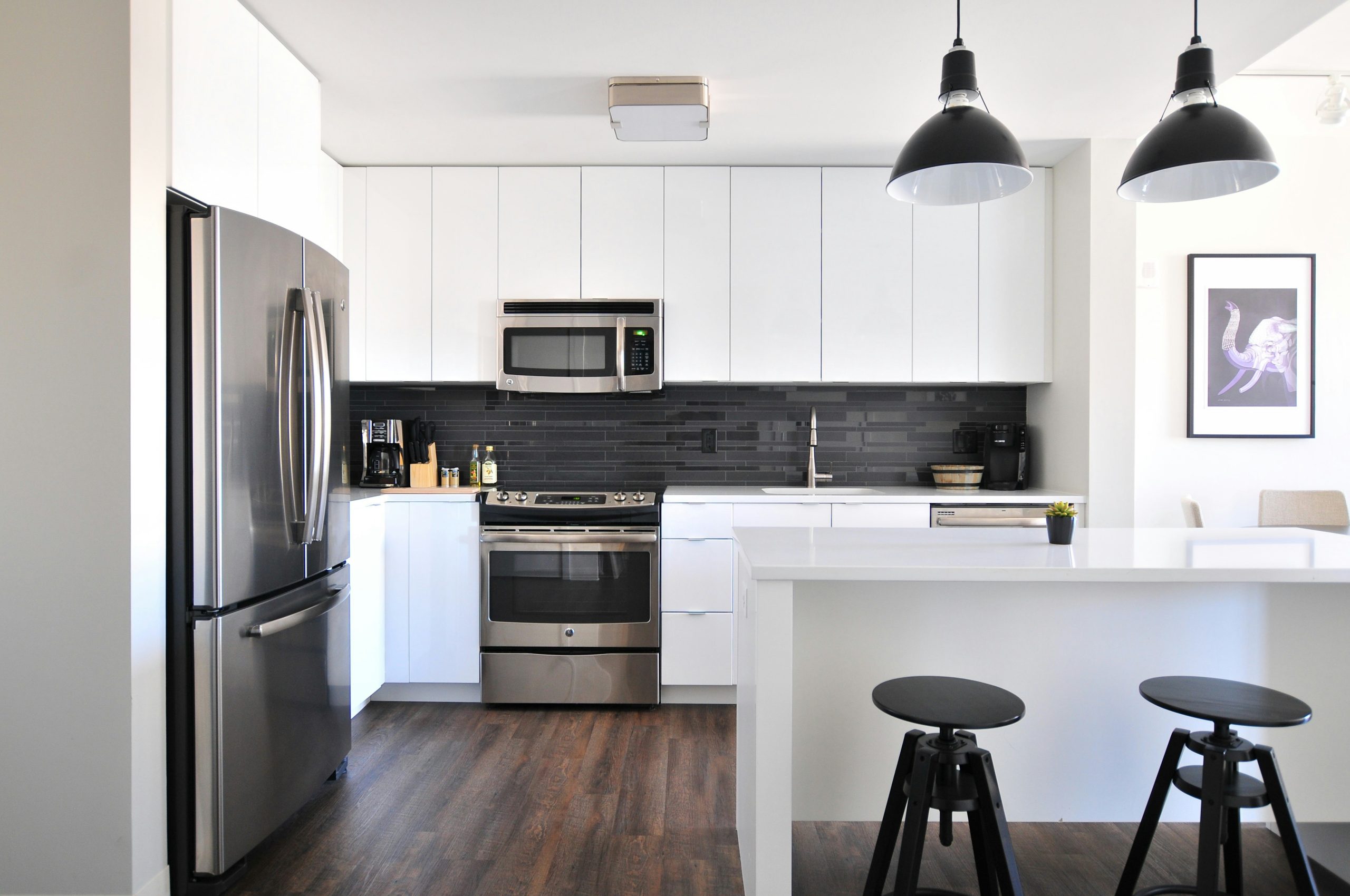 Modern white kitchen with stainless steel appliances, black tile backsplash, pendant lighting, and minimalist island seating.