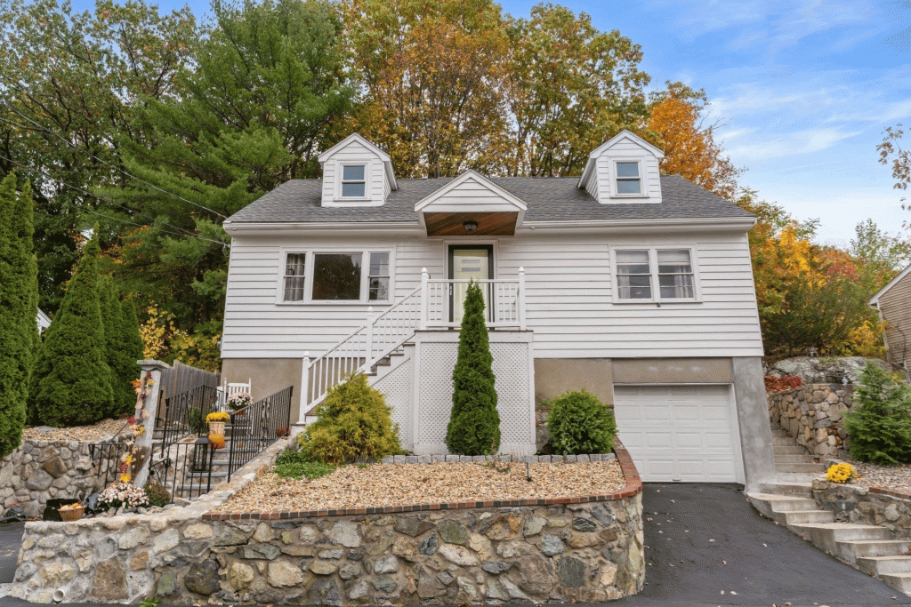 Front exterior view of 43 Maurice Street in Medford, MA — a white Cape-style home with dormers, stone retaining walls, and fall foliage in the background.