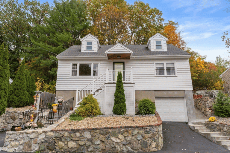 Front exterior view of 43 Maurice Street in Medford, MA — a white Cape-style home with dormers, stone retaining walls, and fall foliage in the background.
