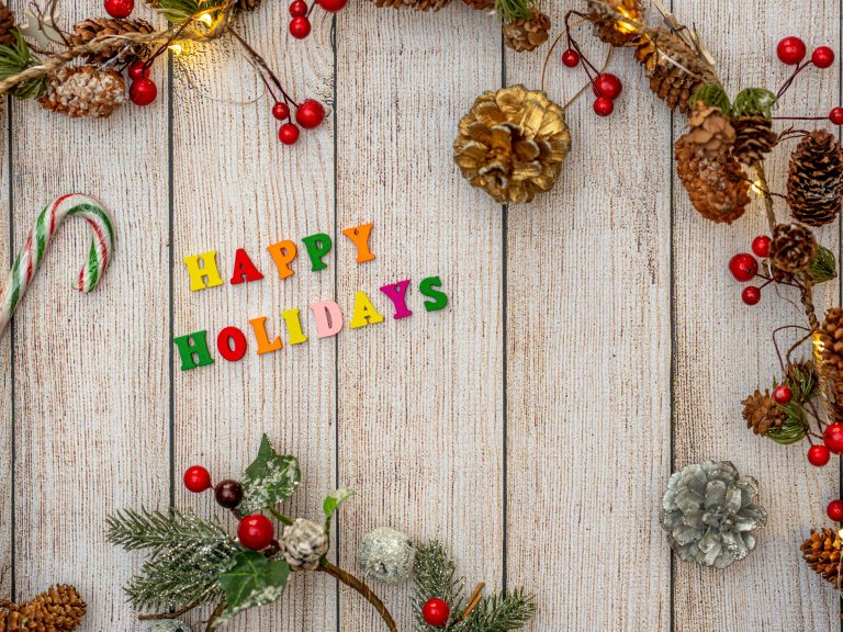 Colorful “Happy Holidays” letters surrounded by pinecones, greenery, berries, and festive decorations on a rustic wooden background